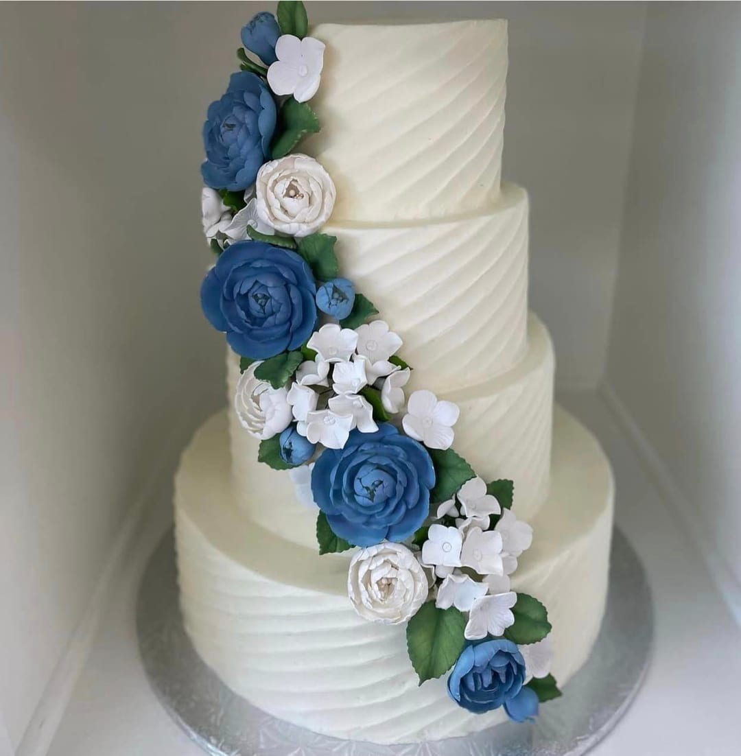 A four-tier white buttercream wedding cake with a cascade of blue and white sugar flowers, showing how each tier can carry its own flavor at Sweet Escape Cake Company in Indianapolis.