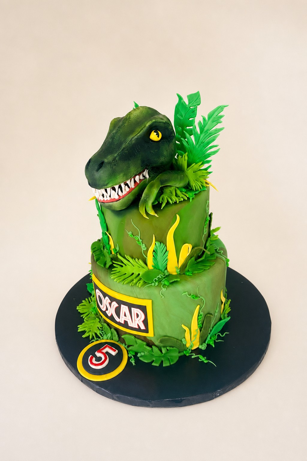 A two-tier sculpted dinosaur birthday cake with a hand-built fondant head and jungle leaves by Sweet Escape Cake Company, the hours of hand work that explain why custom cakes cost more than store bought.