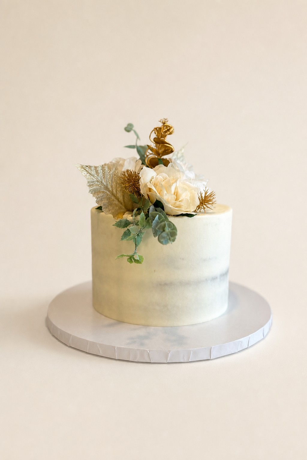 A single tier naked wedding cake with cream flowers and gold leaves showing the exposed cake layers, a rustic wedding cake style by Sweet Escape Cake Company.