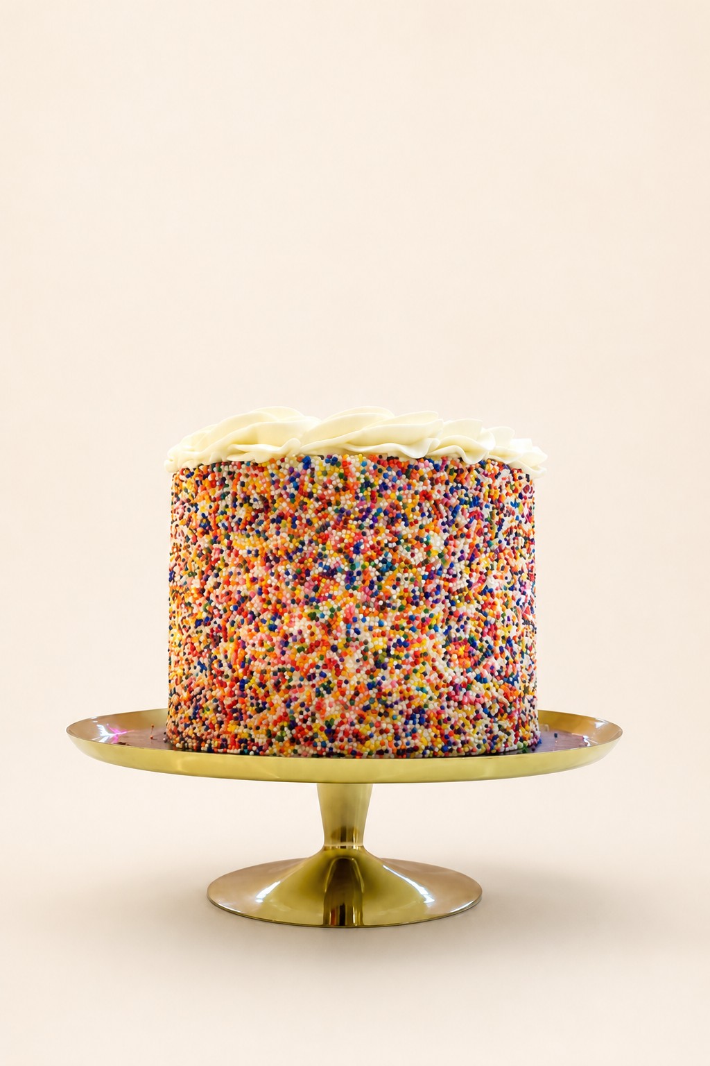 A rainbow sprinkle covered single-tier first birthday party cake with a white buttercream swirl on a gold stand from an Indianapolis bakery.
