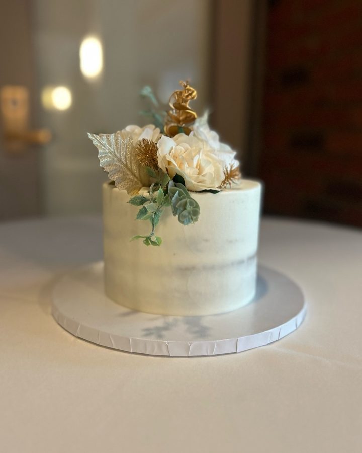 A single-tier round naked cake decorated with cream-colored flowers, shimmering gold leaves, green foliage, and a delicate gold topper, perf