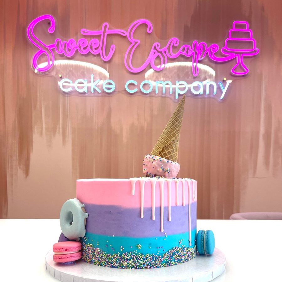A vibrant single-tier ombre buttercream birthday cake featuring pink, purple, and blue colors, adorned with a white drip, colorful sprinkles