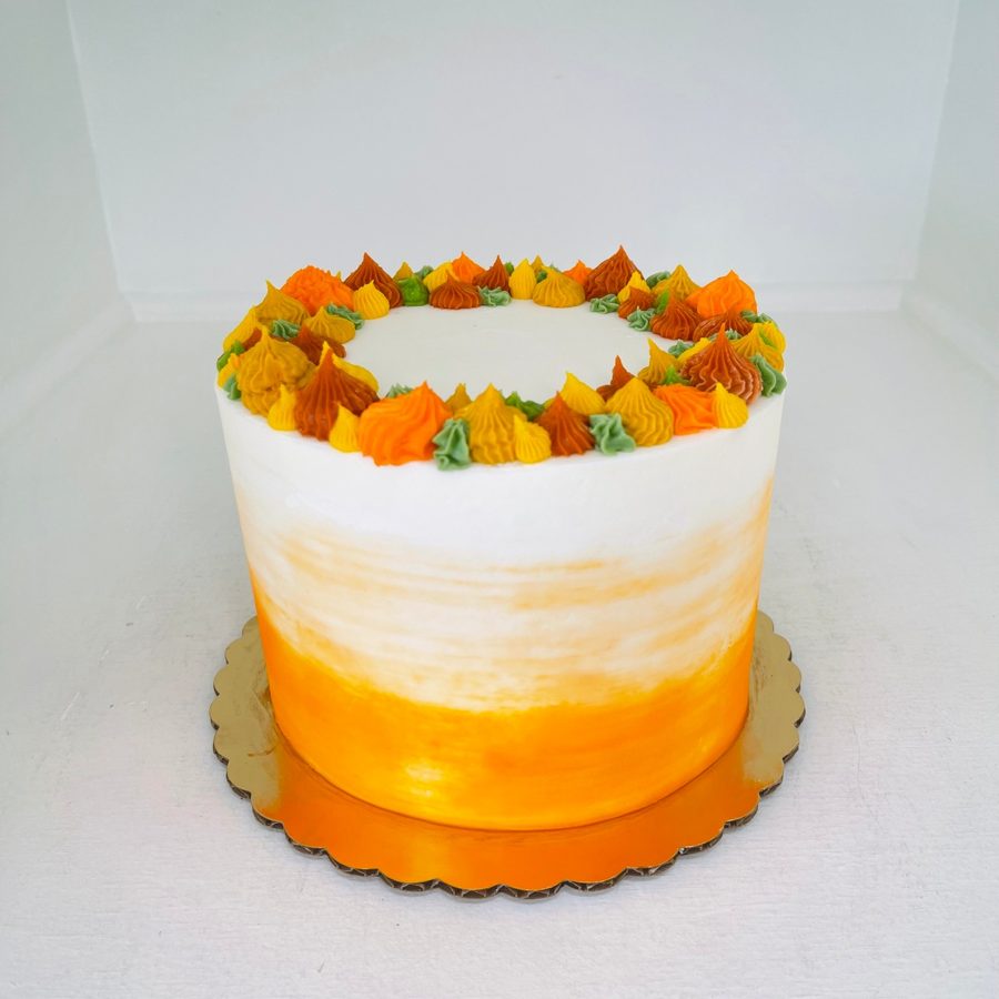A round, single-tier specialty cake featuring a vibrant orange ombre buttercream base and a crown of multi-colored piped frosting swirls in 