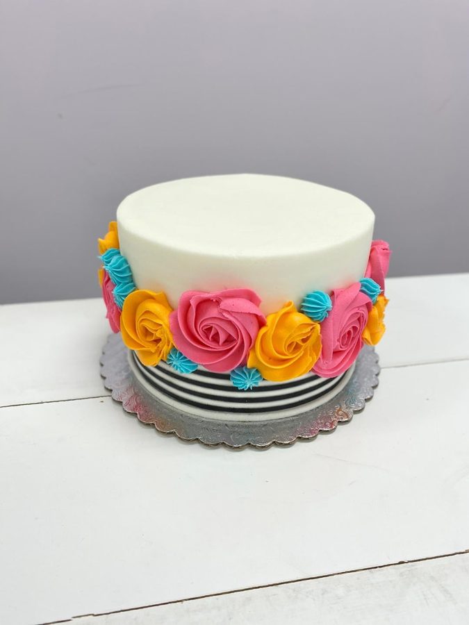 A festive single-tier round white buttercream cake adorned with a vibrant band of piped hot pink, yellow, and turquoise buttercream flowers 
