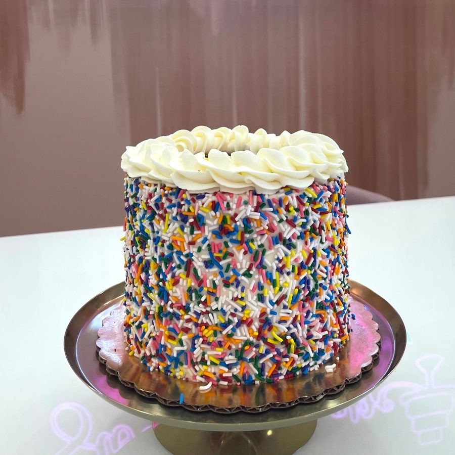 A vibrant round birthday cake featuring white buttercream frosting completely covered in an abundance of colorful sprinkles, topped with a d