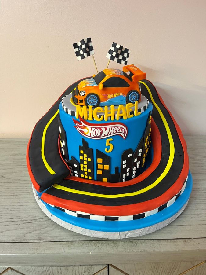 A vibrant Hot Wheels themed two-tier birthday cake for Michael's 5th birthday, featuring an orange race car, a city skyline, racing flags, a