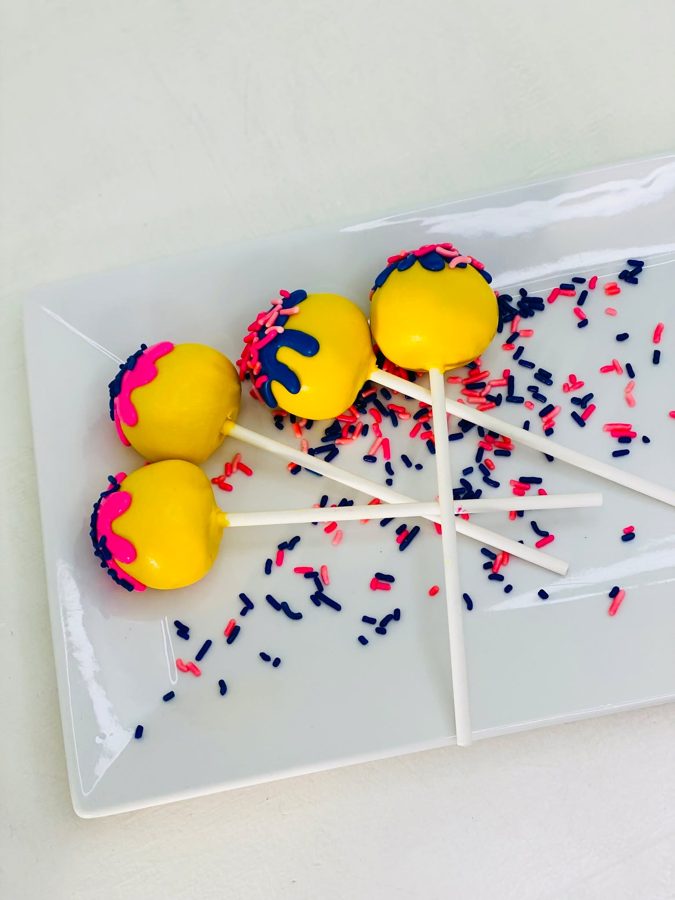 Four bright yellow cake pops decorated with vibrant pink and blue drizzle and matching sprinkles, displayed on a white rectangular plate.