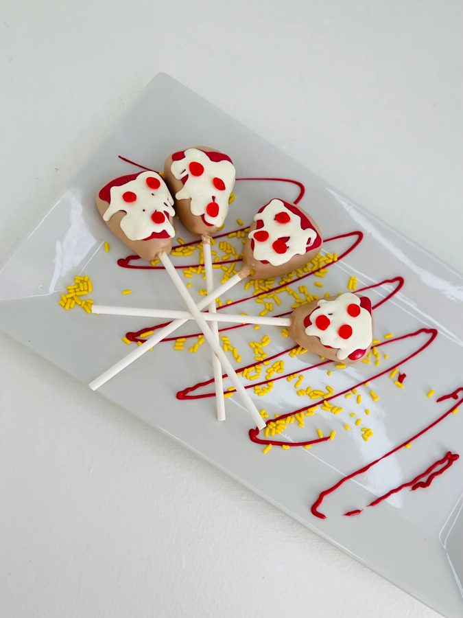 Four pizza-themed cake pops on sticks, decorated with white icing, red pepperoni dots, red drizzle, and yellow sprinkles on a white platter.