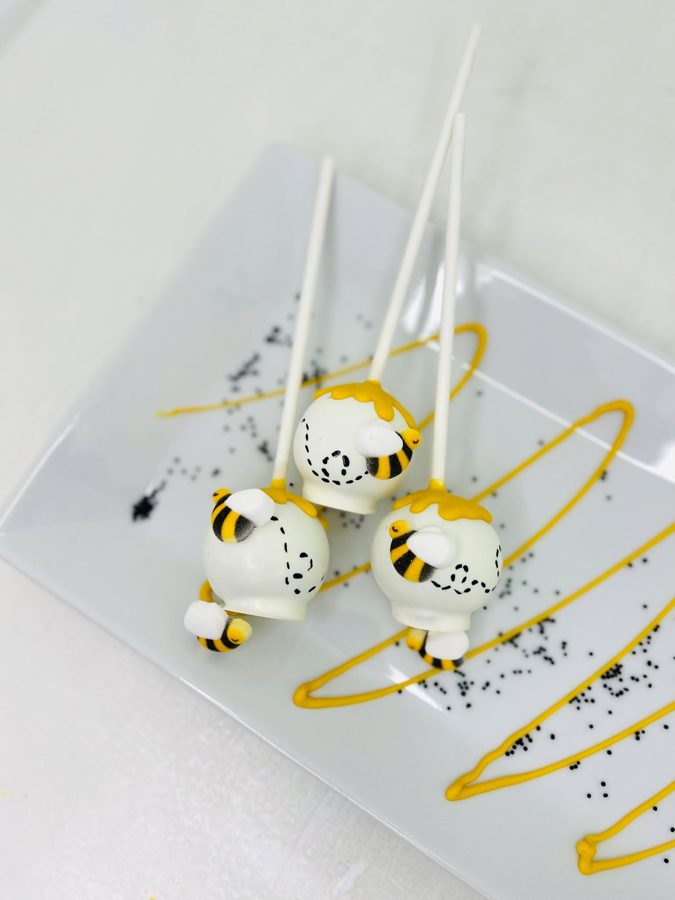 White bee-themed cake pops decorated with yellow drip, black sprinkles, and adorable bumblebee fondant figures.