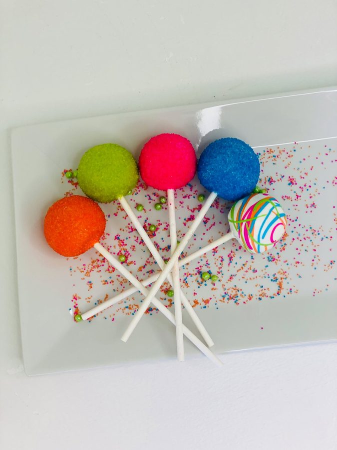 Colorful round cake pops, some coated in vibrant sugar sprinkles (orange, green, pink, blue) and one white cake pop decorated with multicolo