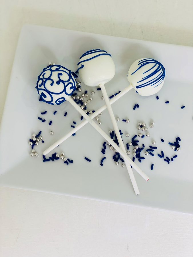 Three white cake pops, two decorated with blue stripes and one with elaborate blue swirl designs, resting on a white plate scattered with na