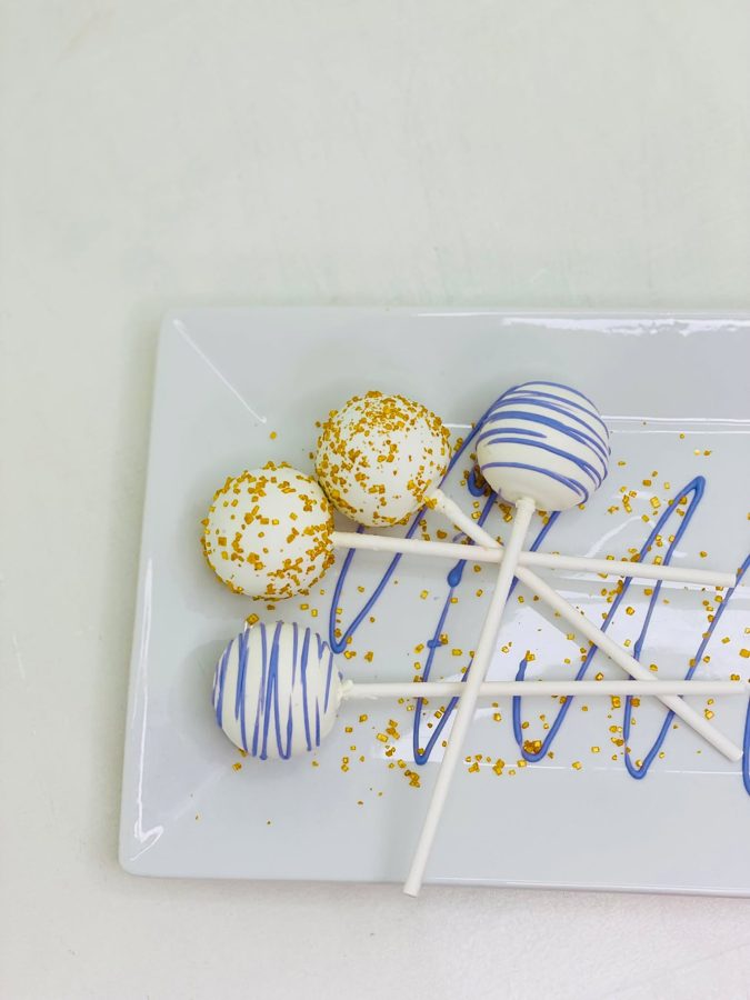 White cake pops decorated with gold sprinkles and blue or purple drizzle, presented on a white rectangular platter.