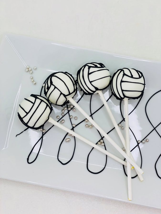 White cake pops decorated with black lines to resemble volleyballs, arranged on a white rectangular plate with scattered silver sprinkles.