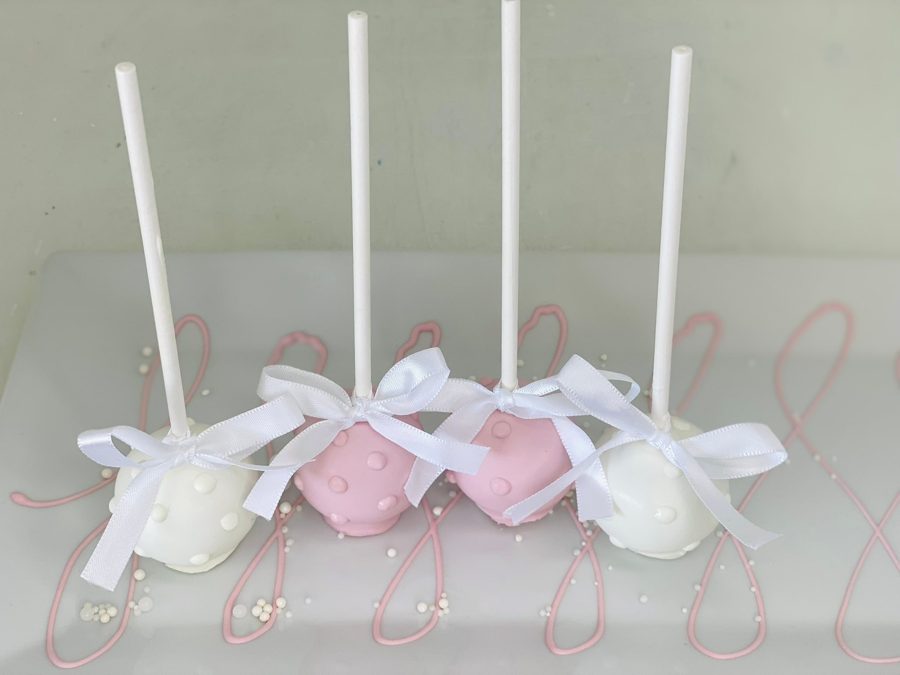 Four elegant cake pops, two white and two pink, each adorned with small raised dots and a delicate white ribbon bow, arranged on a light gre