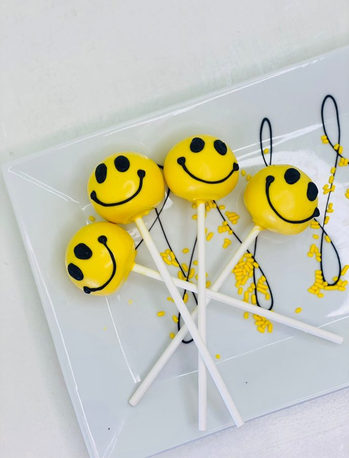 Four cheerful yellow smiley face cake pops with black icing details, presented on a white rectangular plate with yellow sprinkles.