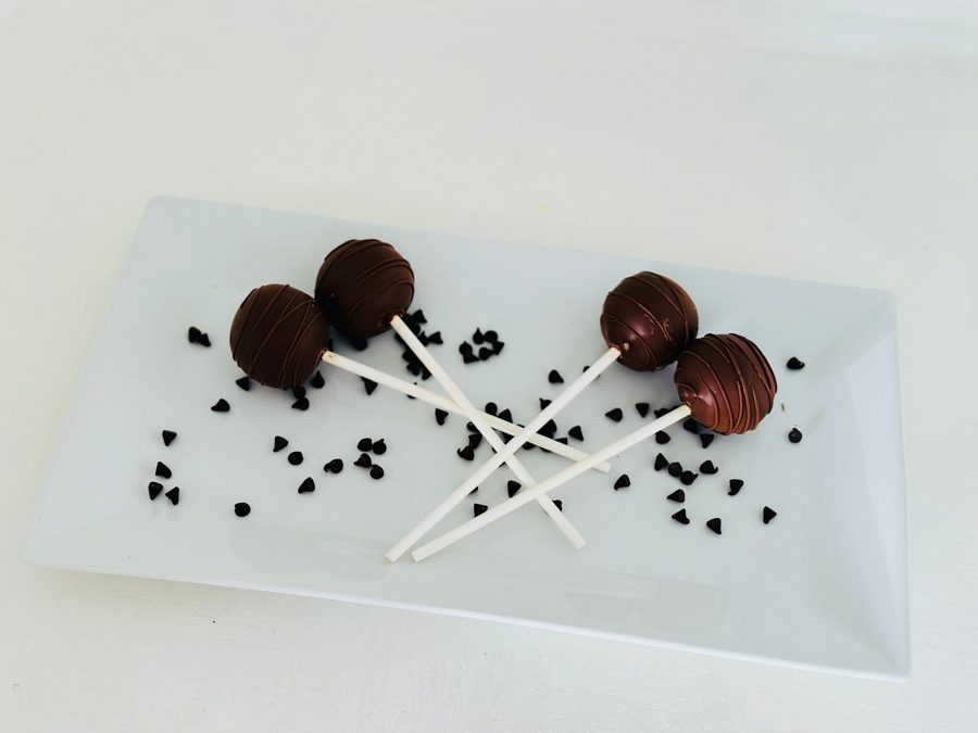 Delicious chocolate cake pops with a delicate chocolate drizzle design, elegantly presented on a white plate with scattered chocolate chips.