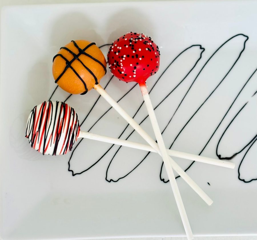 Three assorted cake pops, including a basketball-themed orange cake pop, a red cake pop with black and white sprinkles, and a white cake pop