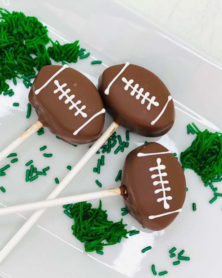 Three chocolate-covered football cake pops decorated with white icing laces and scattered green sprinkles and piped green icing resembling g