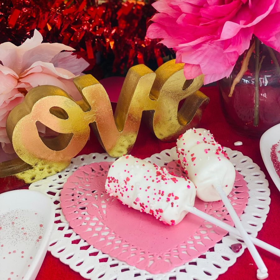White cake pops adorned with red and pink heart-shaped and round sprinkles, presented on heart-shaped doilies with a golden 'LOVE' sign and 