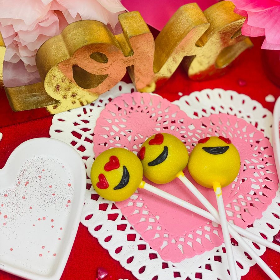 Cheerful yellow emoji cake pops with red heart eyes and black smiles, presented on heart-shaped doilies for a festive Valentine's Day celebr