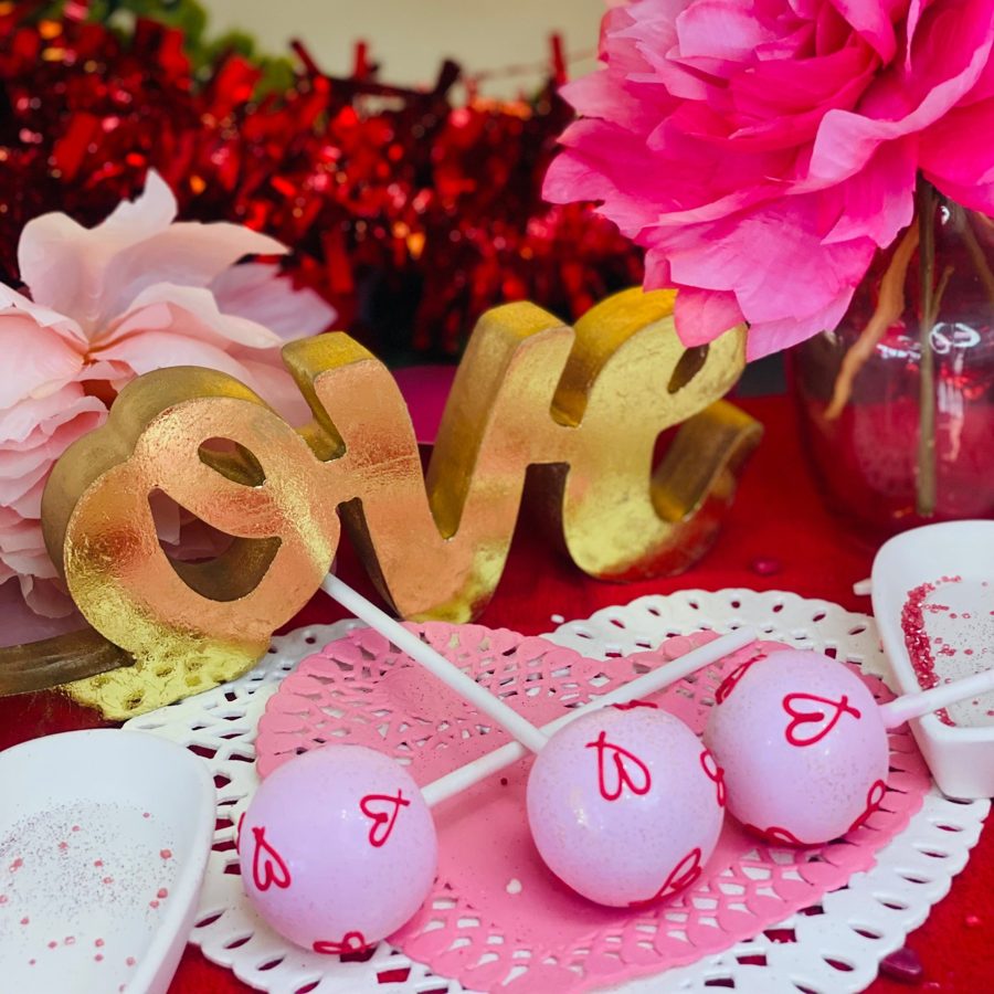 Pink cake pops decorated with red hearts and subtle glitter, displayed on heart-shaped doilies next to a gold 'LOVE' sign, perfect for Valen