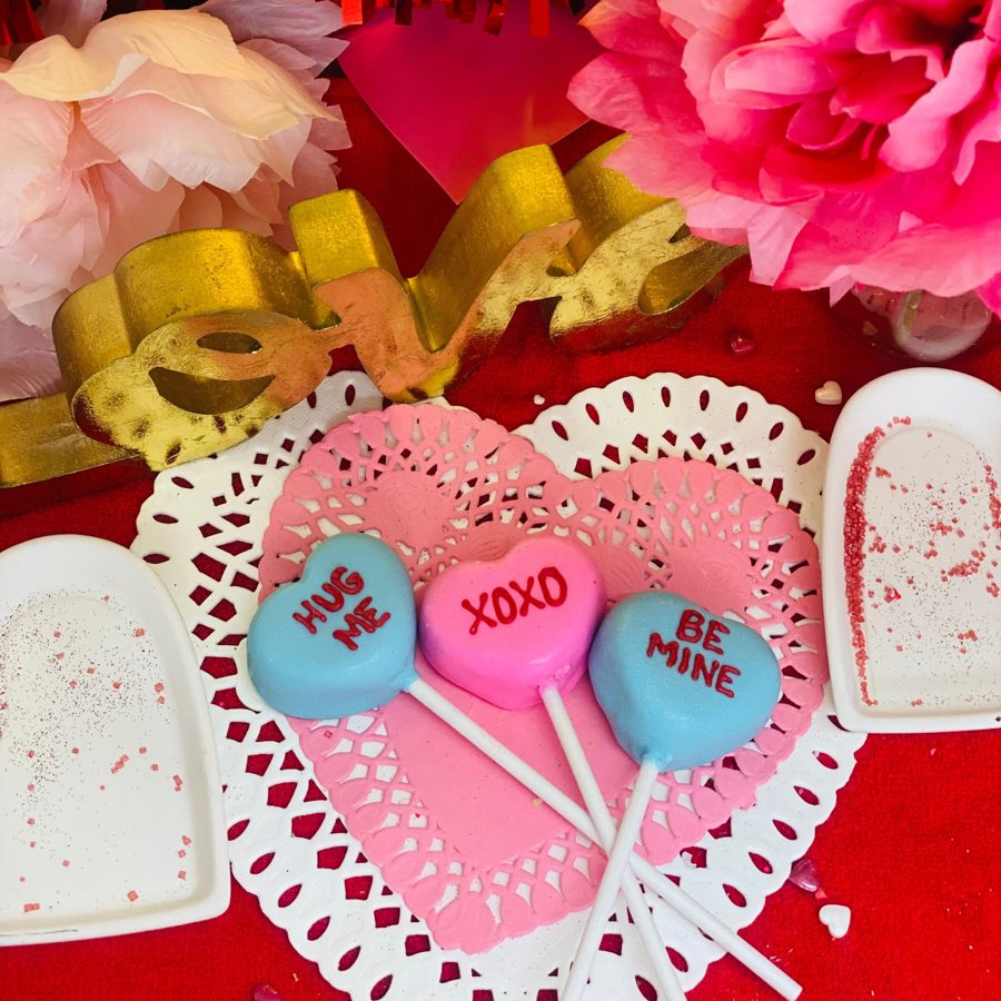 Heart-shaped cake pops in blue and pink with Valentine's Day messages like 'HUG ME', 'XOXO', and 'BE MINE' displayed on lace doilies, surrou