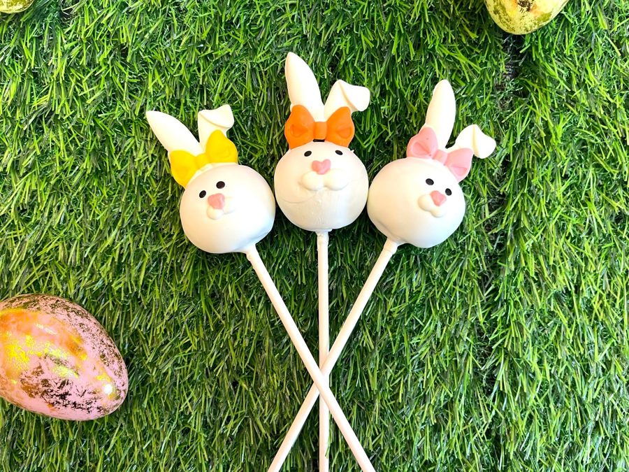 Three cute white bunny cake pops with colorful bows, perfect for an Easter celebration, presented on a vibrant green grass background.