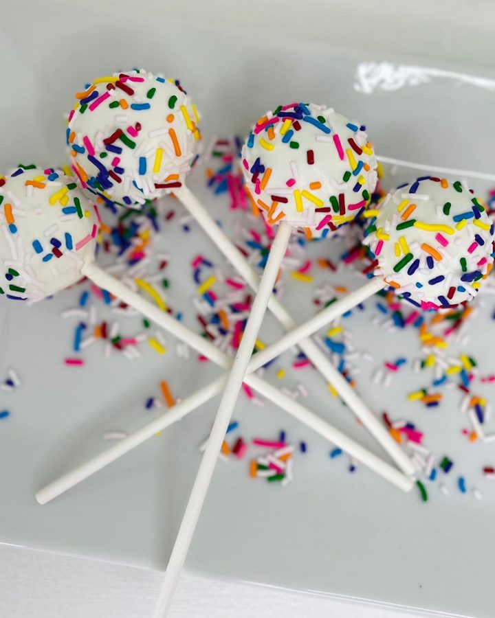 White cake pops coated in colorful rainbow sprinkles, presented on white sticks.