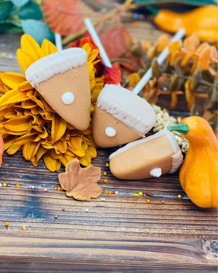 Autumn-themed cake pops or dessert treats shaped like pumpkin pie slices and acorns, decorated with cream-colored icing and sprinkles, prese