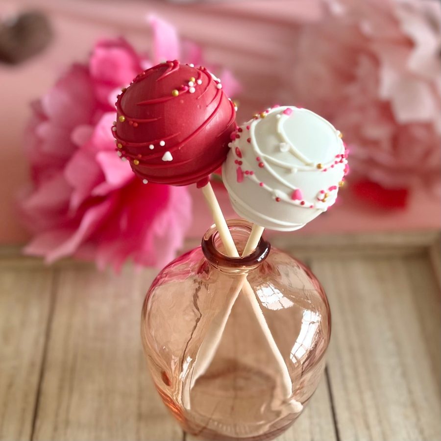 Two elegant red and white cake pops adorned with gold and pink sprinkles, including tiny heart shapes, presented in a small vase.