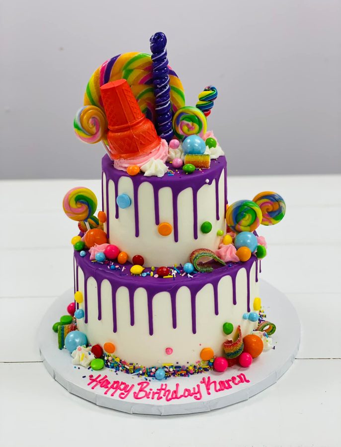 A two-tiered white birthday cake with purple drip icing, generously decorated with an assortment of colorful candies, lollipops, and sprinkl