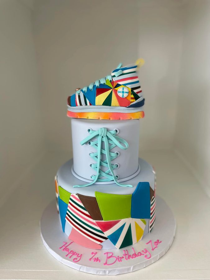 A vibrant two-tier round birthday cake featuring a colorful patchwork design, a detailed sneaker figurine on top, and light blue fondant lac