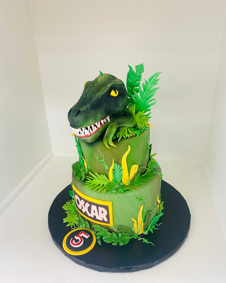 A two-tiered green fondant dinosaur birthday cake featuring a sculpted dinosaur head, jungle leaves, and personalized with the name OSCAR an