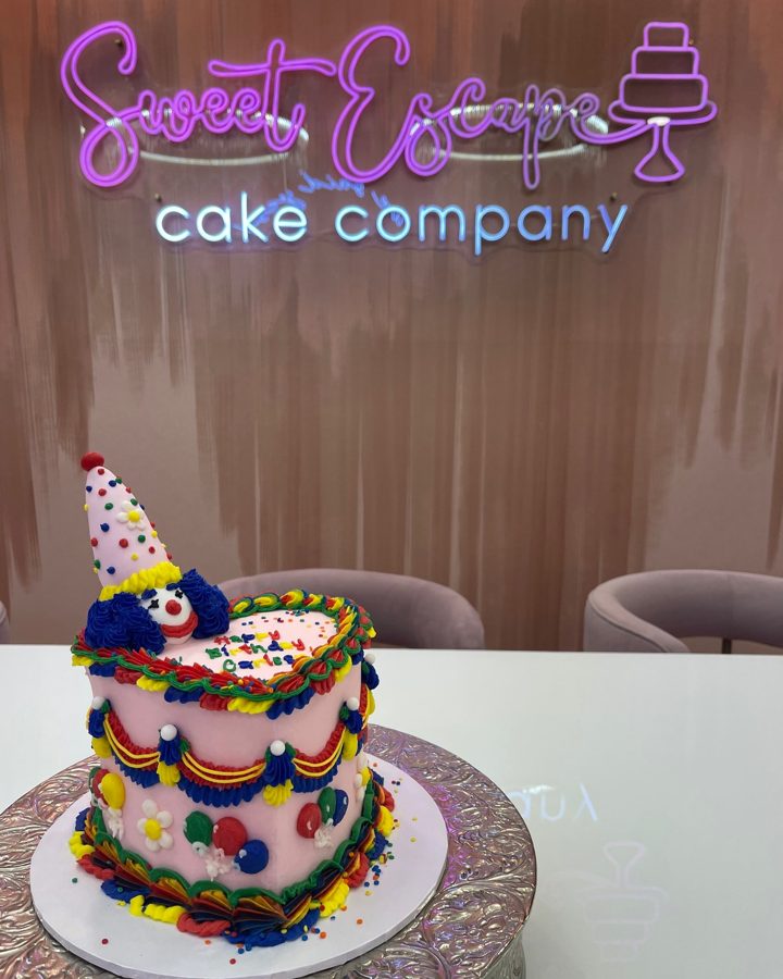 A vibrant pink buttercream birthday cake featuring a cheerful clown topper with a polka-dot party hat, adorned with colorful piped garlands,