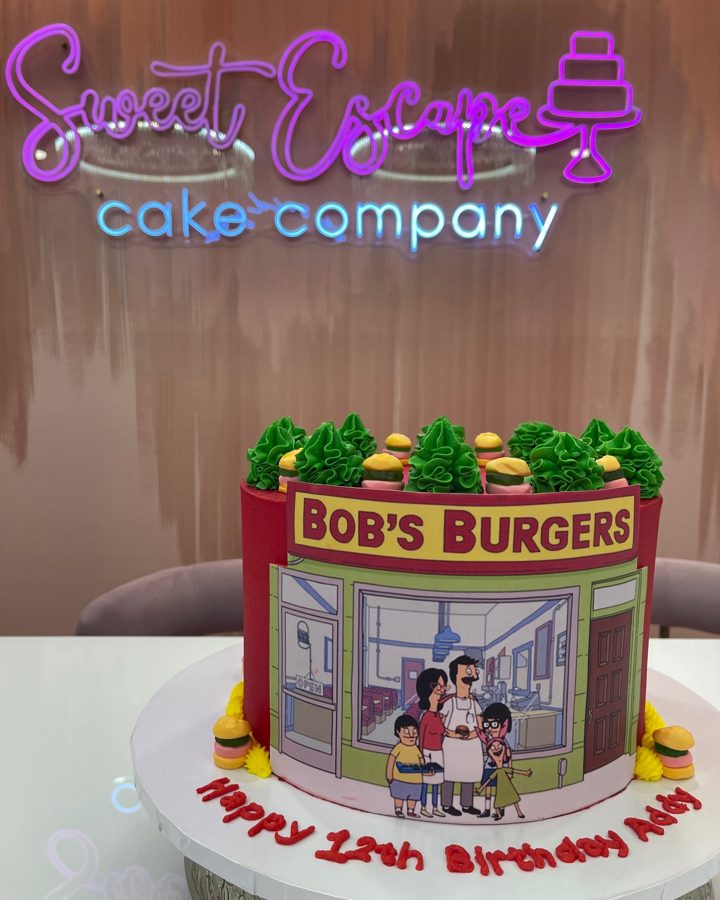 A vibrant red Bob's Burgers themed birthday cake featuring an edible image of the restaurant and characters, topped with green frosting tree