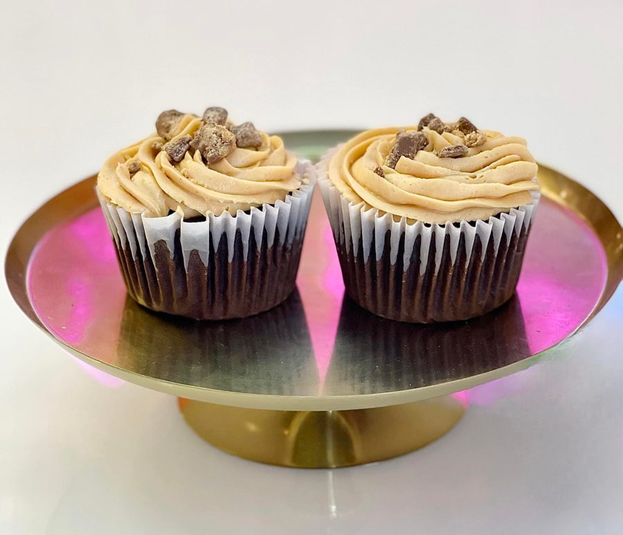 Two chocolate cupcakes with tan-colored buttercream frosting, topped with crunchy brown candy pieces, presented on a reflective gold display