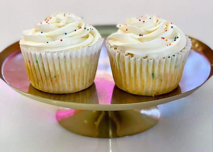 Two classic funfetti cupcakes with fluffy white buttercream frosting and colorful sprinkles, presented on a shiny gold cake stand.