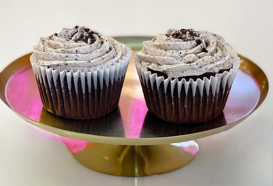 Two decadent chocolate cupcakes topped with creamy cookies and cream frosting and cookie crumb sprinkles, elegantly displayed on a gold dess