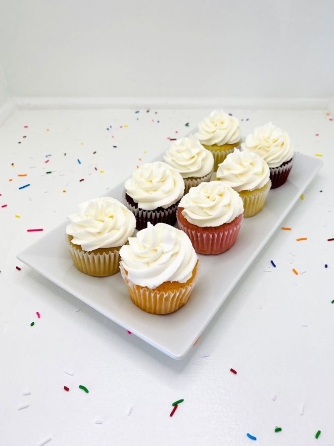 Assorted cupcakes with white buttercream frosting and colorful wrappers, displayed on a white plate surrounded by scattered colorful sprinkl