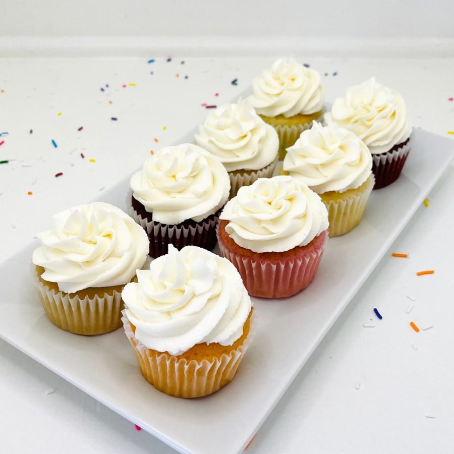 A set of assorted mini cupcakes featuring white swirled buttercream frosting on various cake flavors, with colorful sprinkles scattered on a