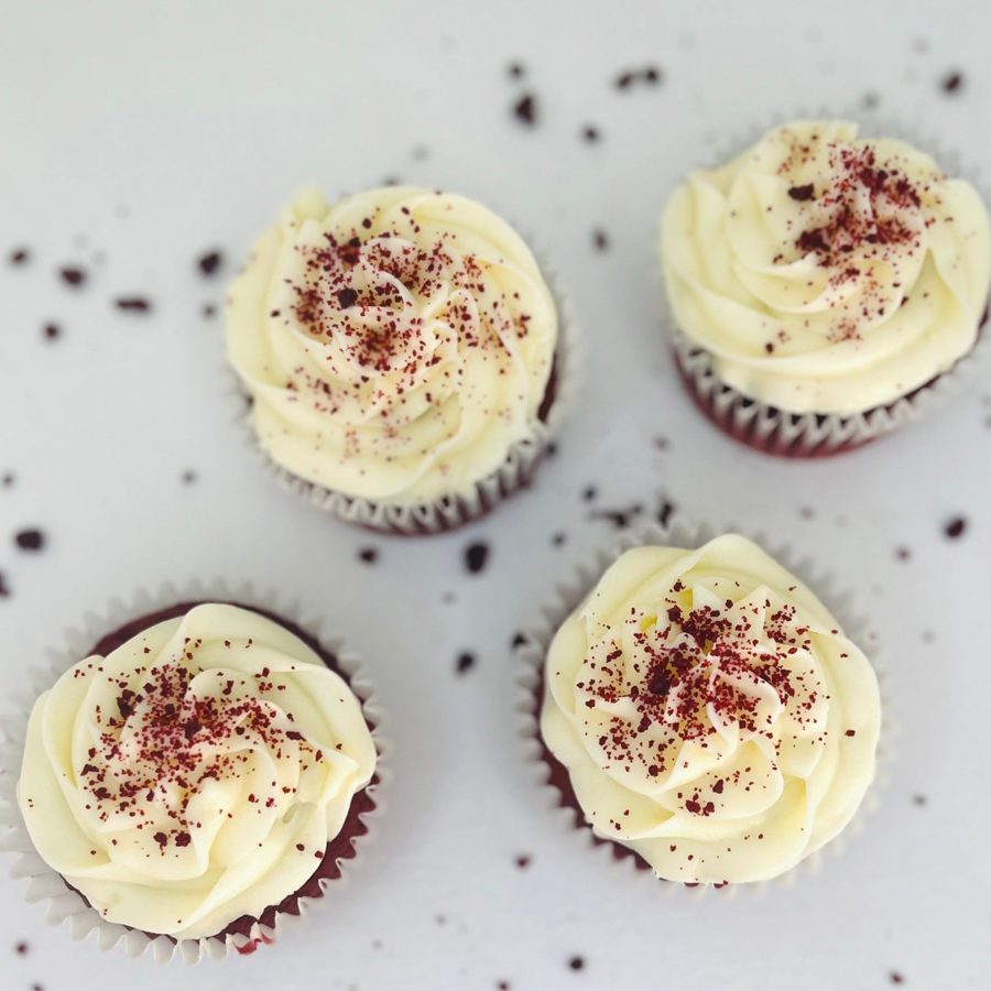 Four beautifully decorated red velvet cupcakes with creamy white frosting and red crumb sprinkles, presented on a minimalist white backgroun