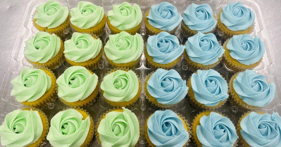 A clear tray of cupcakes featuring light green and light blue rose-shaped buttercream frosting, perfect for a baby shower or gender reveal c