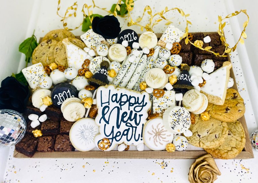 A festive New Year's Eve dessert board featuring an assortment of decorated cookies, brownies, macarons, chocolate-covered pretzels, and mar