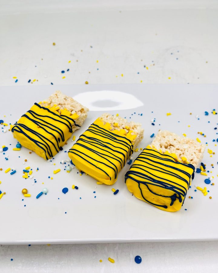 Three rectangular Rice Krispie treats partially dipped in bright yellow candy coating, decorated with thin blue stripes, and surrounded by c
