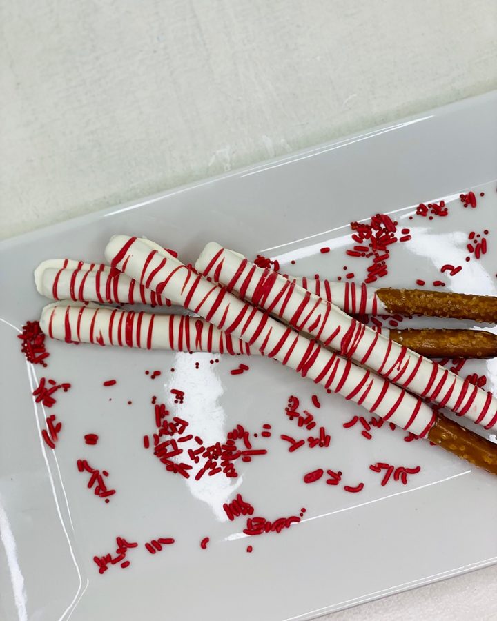 White chocolate-dipped pretzel rods with festive red stripes and scattered red sprinkles, presented on a white serving platter.