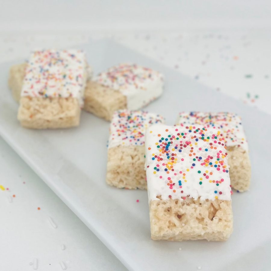 Rectangular Rice Krispie treats half-dipped in a white coating and generously topped with colorful sprinkles, presented on a white serving p