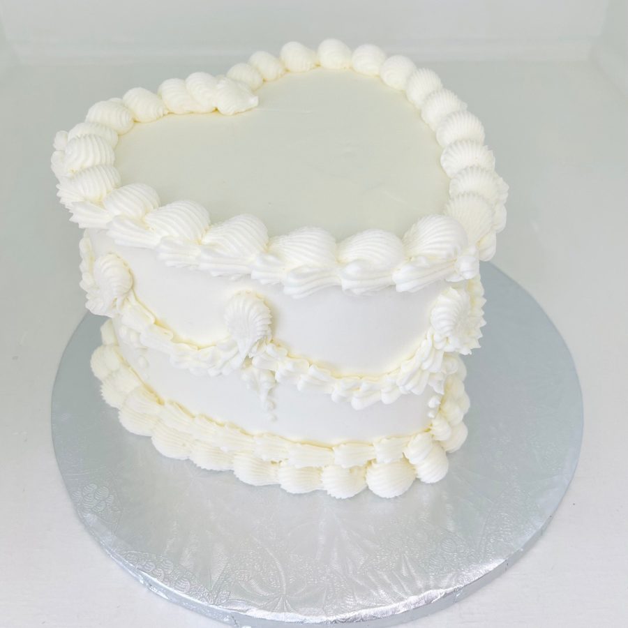 An elegant white heart-shaped cake adorned with intricate buttercream piping, featuring shell borders on the top and bottom edges, and ruffl
