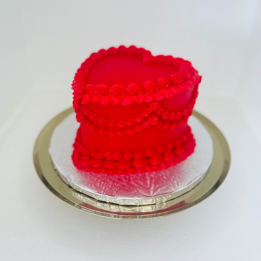 A vibrant red heart-shaped cake featuring intricate piped buttercream ruffles and swirls along the top and sides, presented on a silver cake
