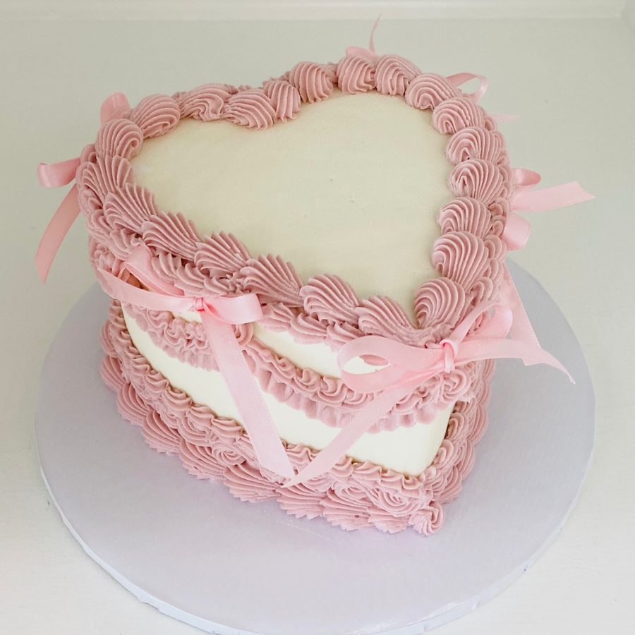 An elegant heart-shaped cake featuring a smooth cream finish, delicate pink ruffled buttercream piping around the edges, and decorative pink