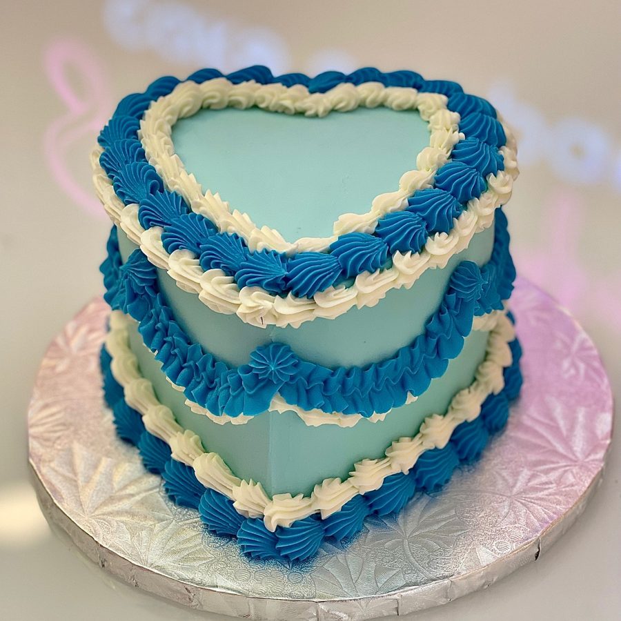 A elegant heart-shaped light blue buttercream cake decorated with intricate white and dark blue piped scrollwork and shell borders.